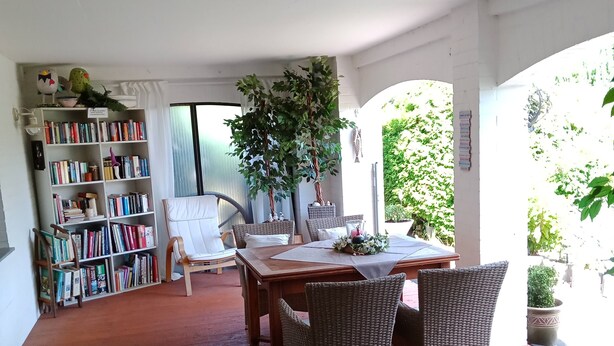 Una terraza de madera con mesa de comedor, sillas de mimbre, una estantería y una planta grande en maceta.