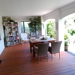 Une terrasse en bois avec une table à manger, des chaises en osier, une bibliothèque et une grande plante en pot.