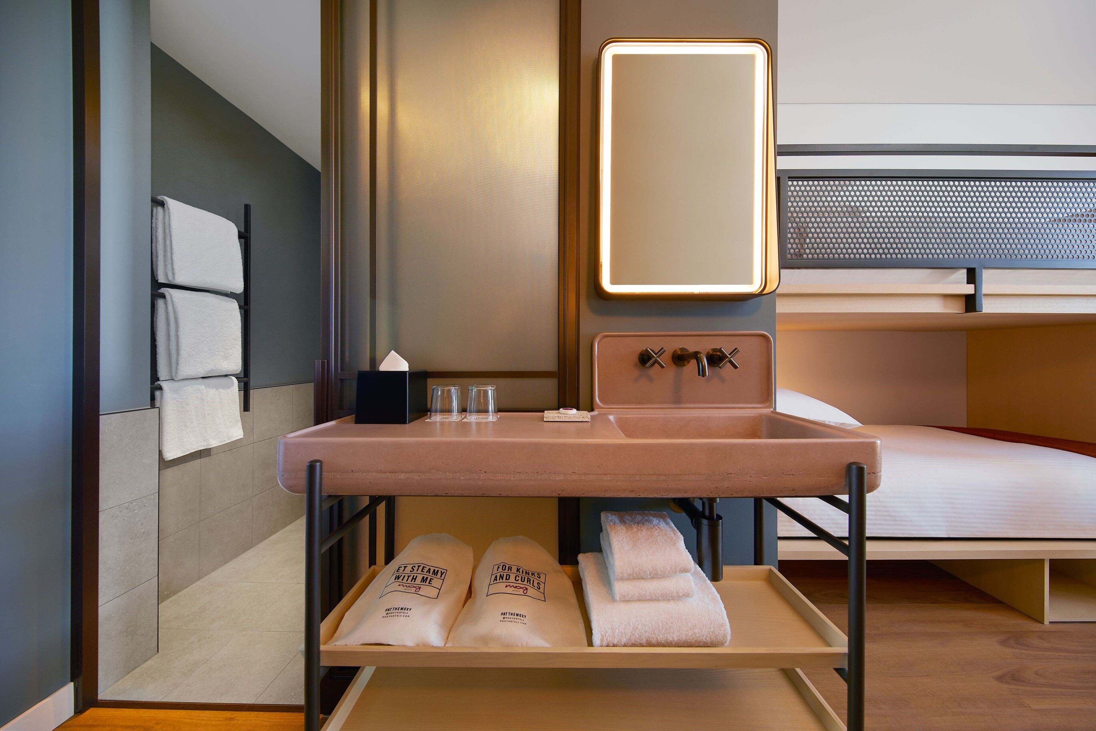 Una habitación de hotel moderna con suelo de madera, una cama con cabecero, un lavabo con espejo, y una estantería con toallas y artículos de aseo.