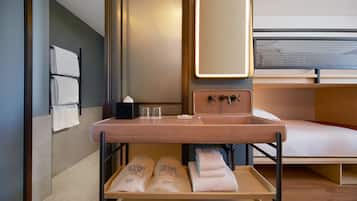 Una habitación de hotel moderna con suelo de madera, una cama con cabecero, un lavabo con espejo, y una estantería con toallas y artículos de aseo.