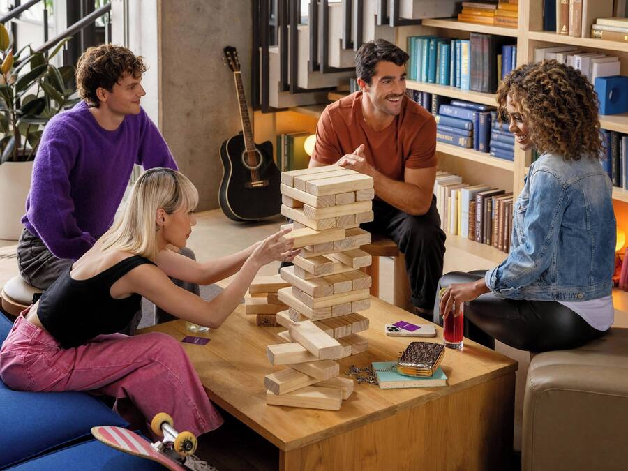 Un grupo de personas jugando un juego de mesa en una habitación acogedora y bien iluminada, con estanterías de libros y una guitarra al fondo.