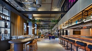 A modern bar area with a long counter and multiple stools, a seating area with tables and chairs, and a visible staircase in the background. - Moxy Sydney Airport (Mascot)
