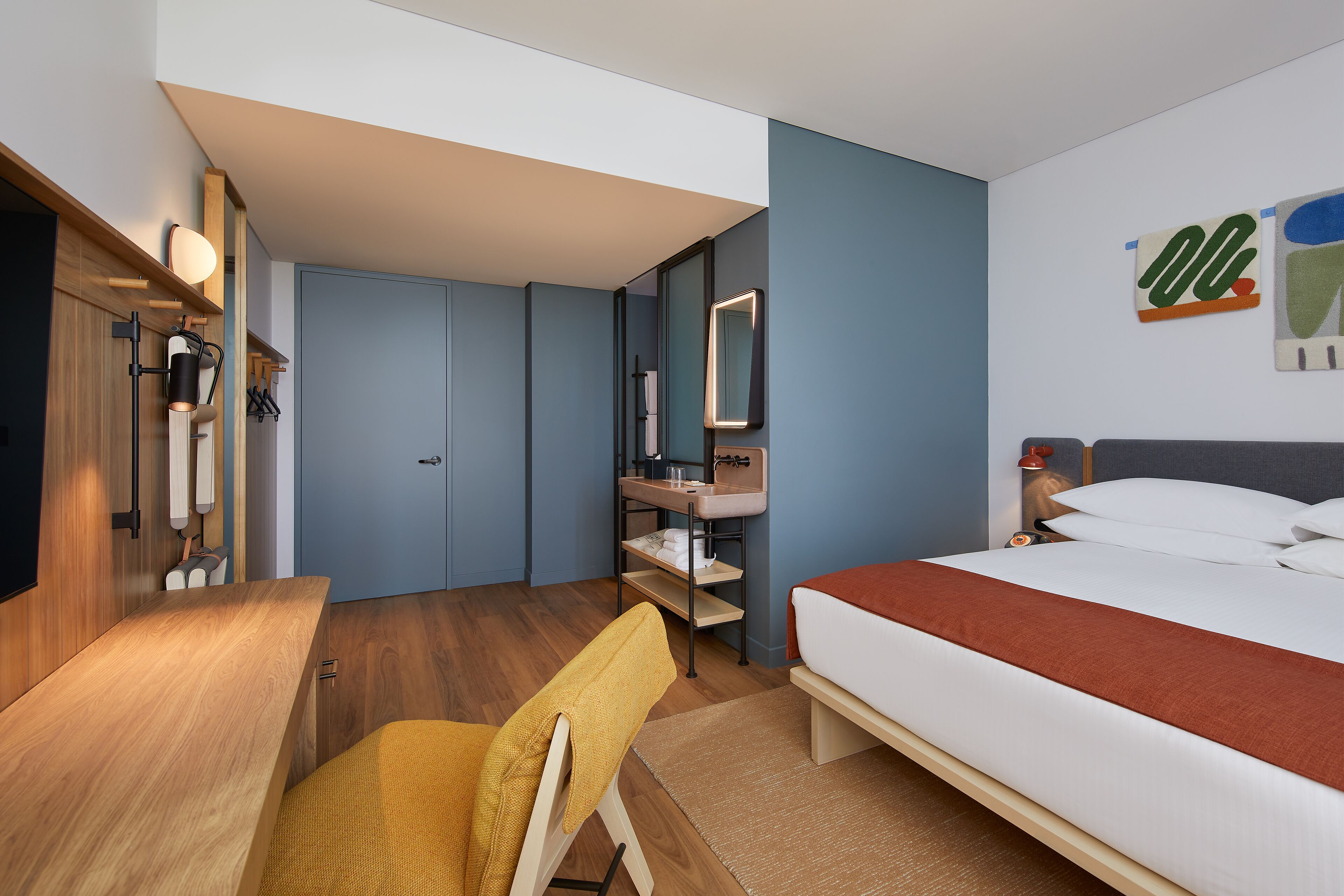 A modern hotel room with a wooden desk, a bed with a red blanket, a bathroom with a mirror, and a wall art piece.