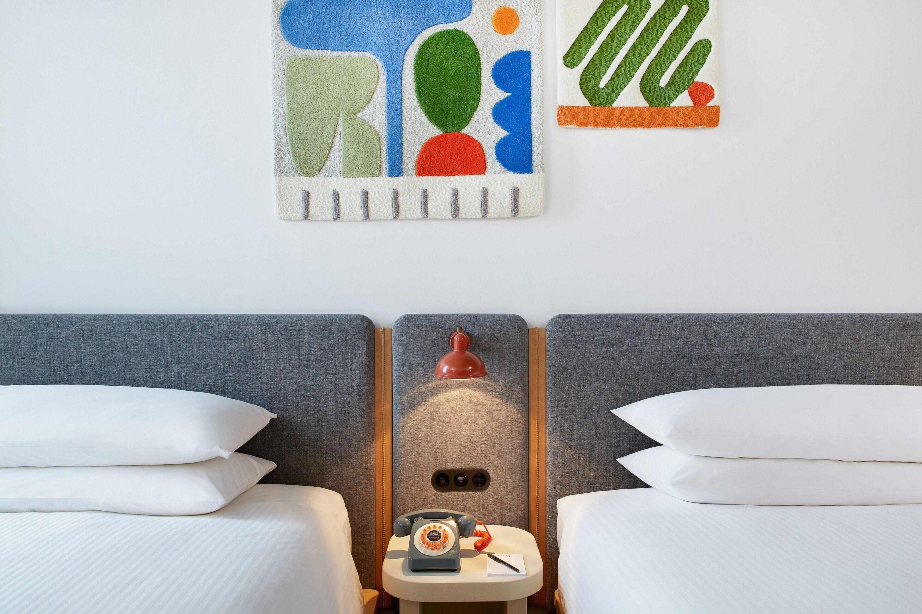 A hotel room with two beds, a small table with a telephone, and a wall-mounted lamp.