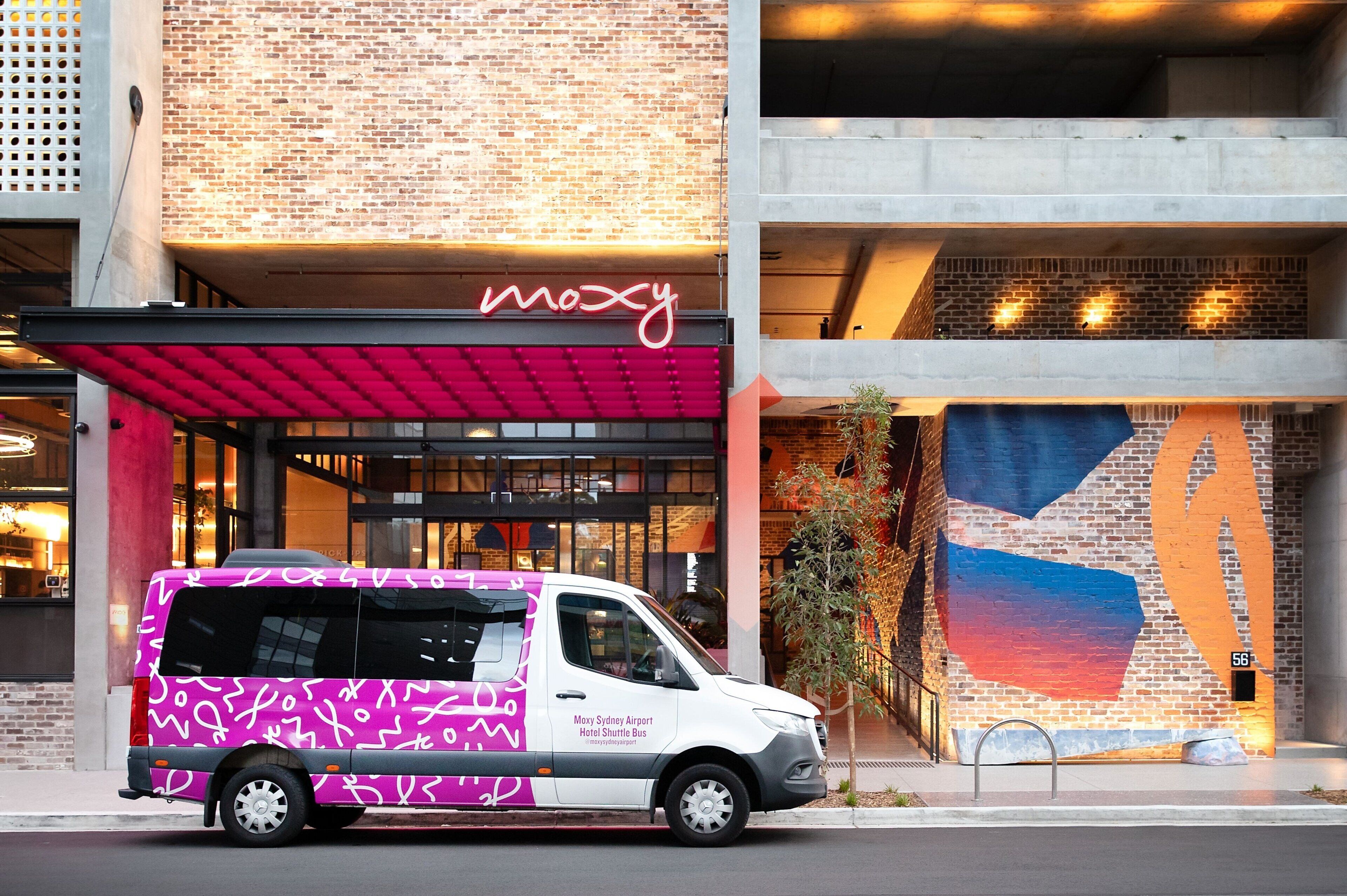 A white shuttle bus with pink and black patterns parked in front of a building with a brick facade and a large, colorful mural.