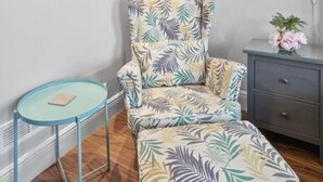 Ein Zimmer mit einem gemusterten Sessel, einem kleinen runden Tisch, einem gerahmten Kunstwerk an der Wand und einem Nachttisch mit einer Lampe.