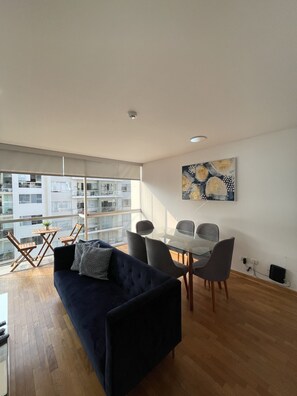 Un salon moderne avec un canapé bleu foncé, une table à manger en verre entourée de chaises et une grande fenêtre donnant sur un immeuble doté de balcons.