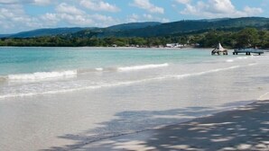 A beach with clear turquoise water, white sand, and a backdrop of green hills under a partly cloudy sky.