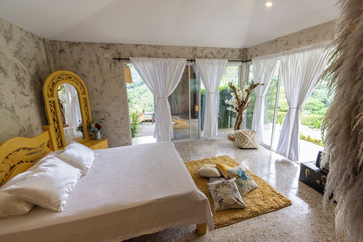 A bedroom with a large bed, a mirror, and a view of greenery through the windows.