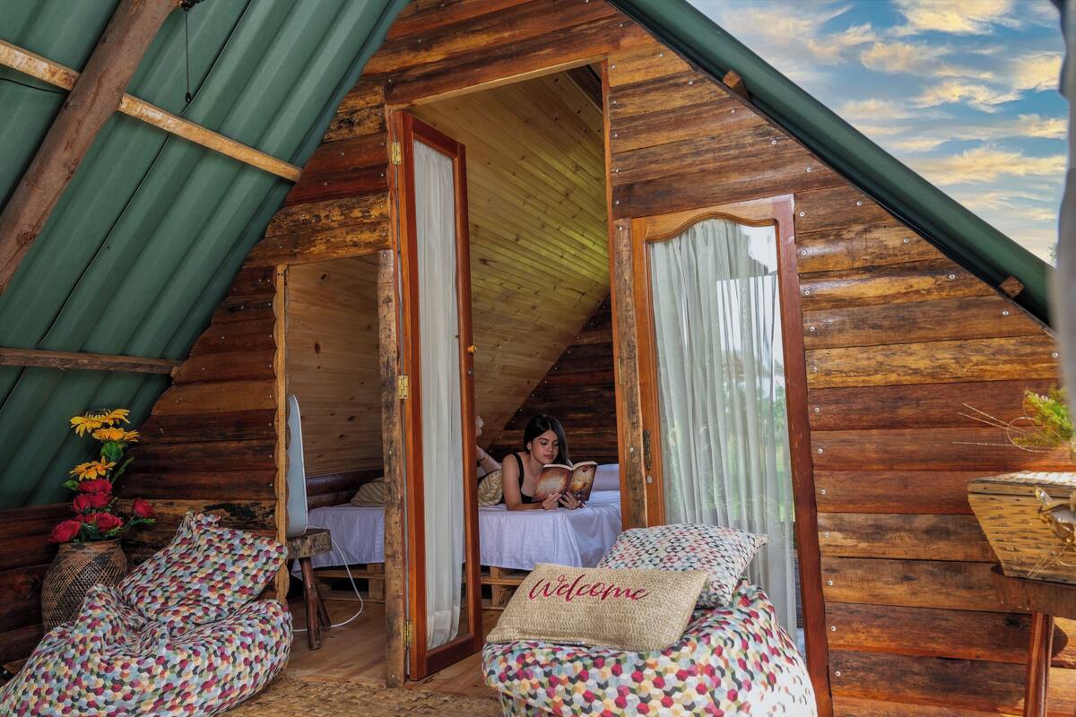 A cozy wooden cabin with a sloped roof, a bed with a 'Welcome' pillow, and a small table with a vase of flowers.