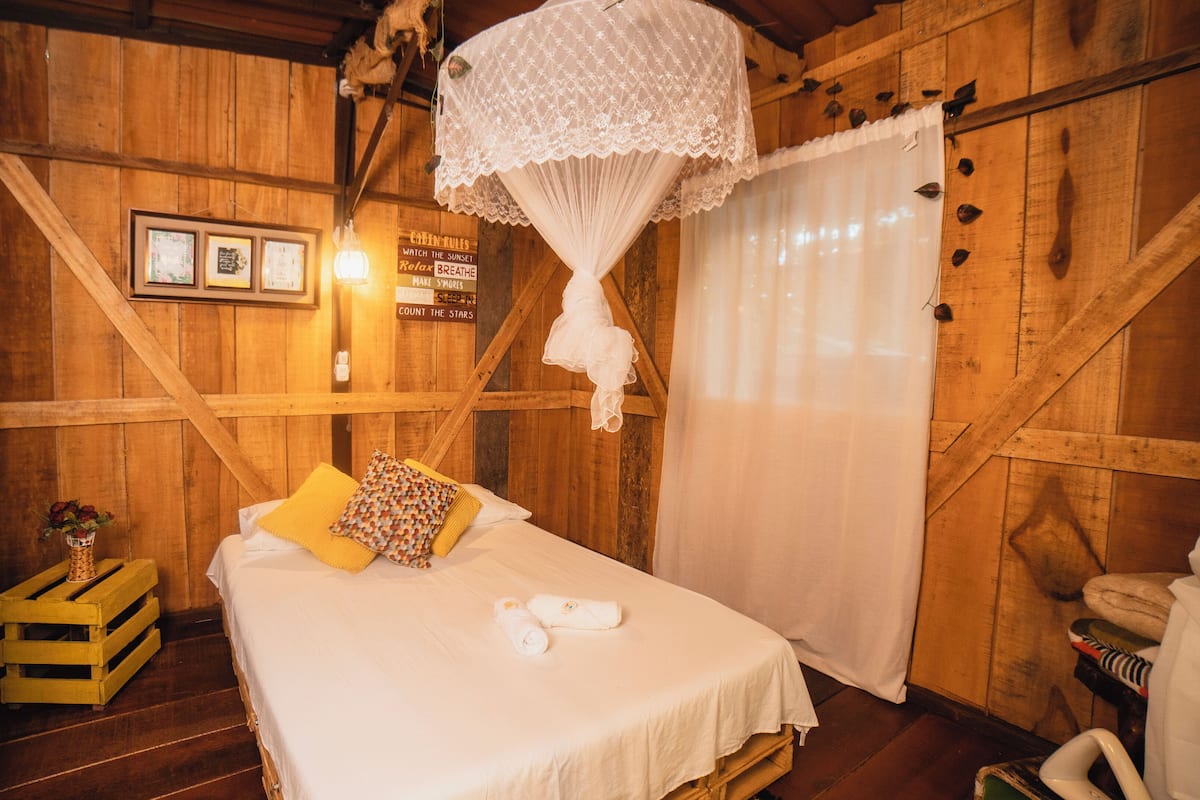 A wooden cabin room with a bed, a nightstand, a small table with a vase of flowers, and a sign with cabin rules.