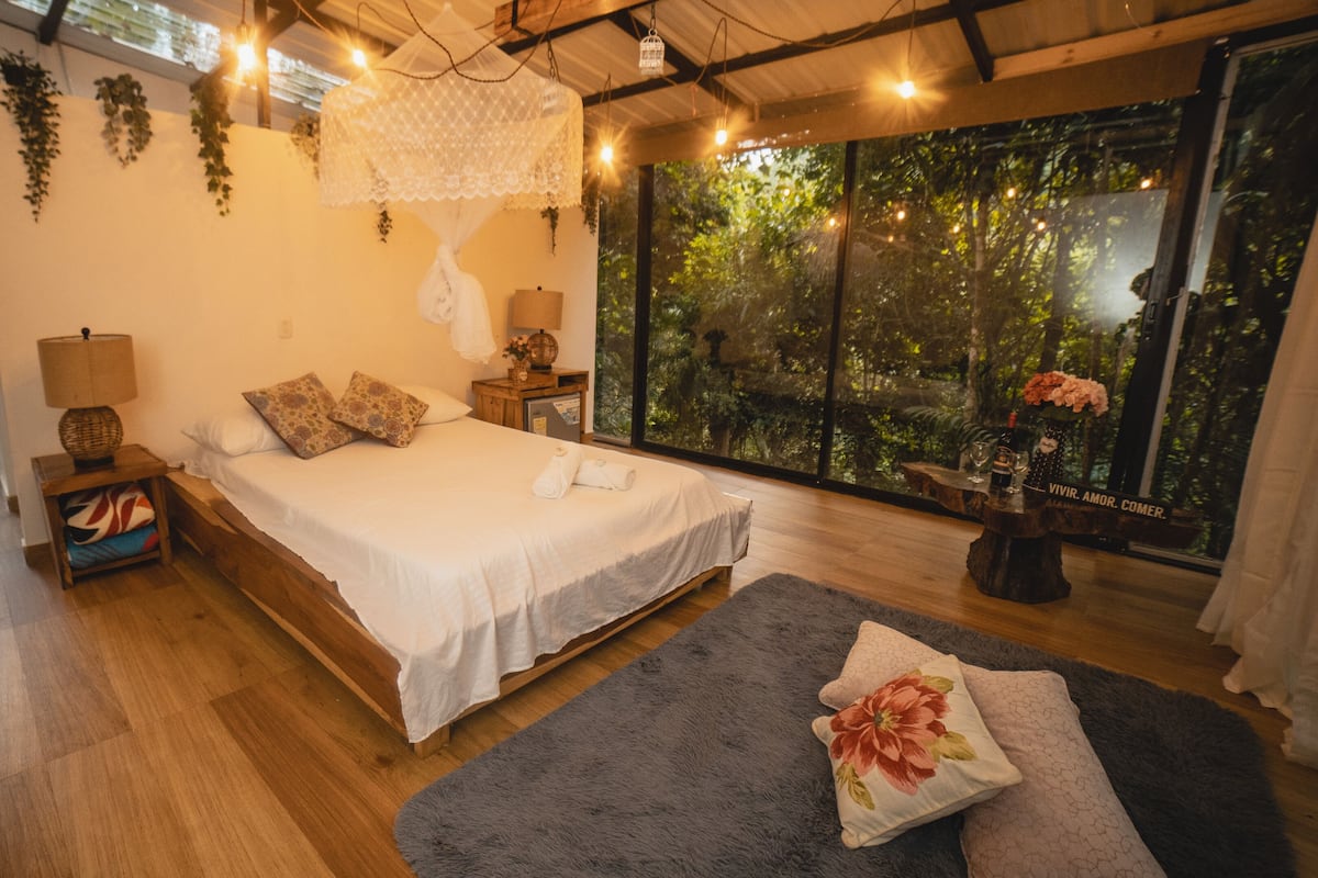 A bedroom with a large bed, bedside tables, a nightstand, a window with a view of trees, and a balcony.