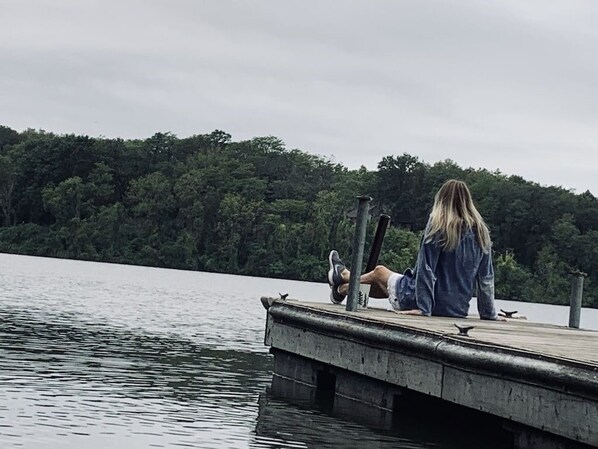 A person sitting on a wooden dock extending into a lake, surrounded by dense forest.