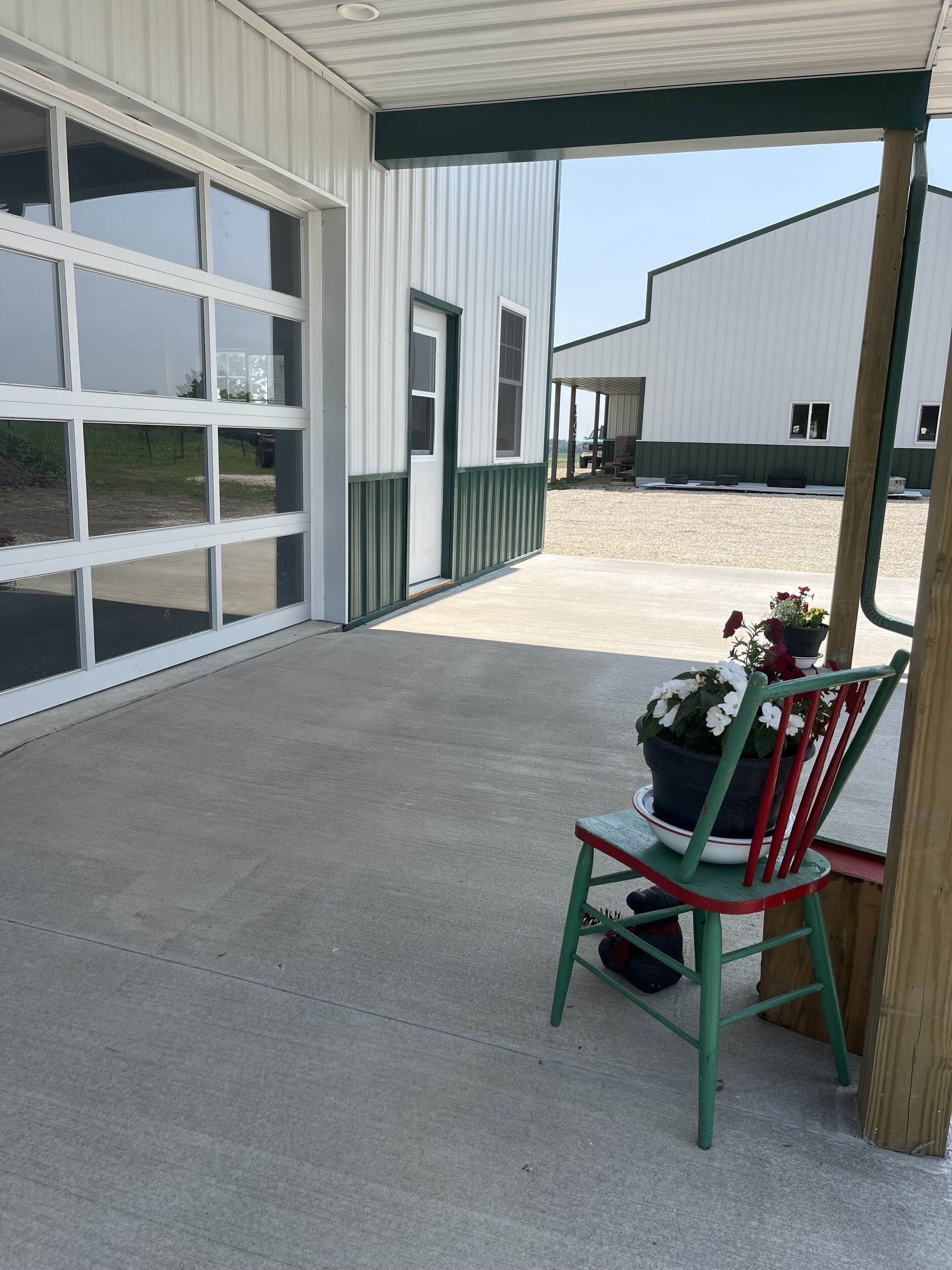 Een overdekt buitengebied met een betonnen vloer, een groene stoel met een plant in pot, en een gebouw met grote ramen en een garage.