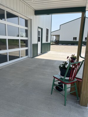 A covered outdoor area with a concrete floor, a green chair with a potted plant, and a building with large windows and a garage door.