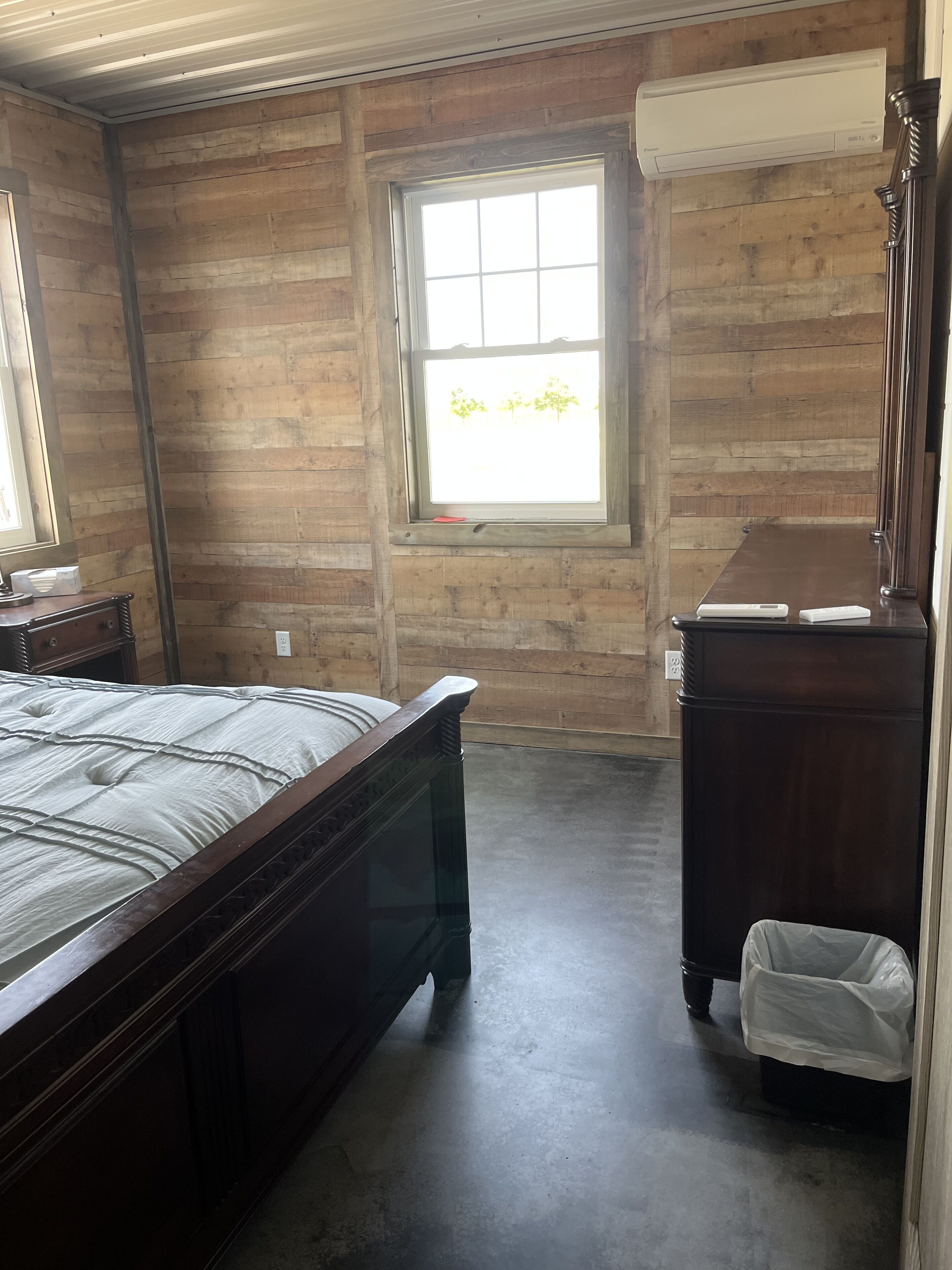 A room with a wooden floor, a bed, a dresser, a window, and a trash bin.