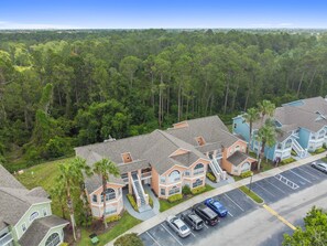 A residential area with multiple houses, a parking lot, and a dense forest in the background.