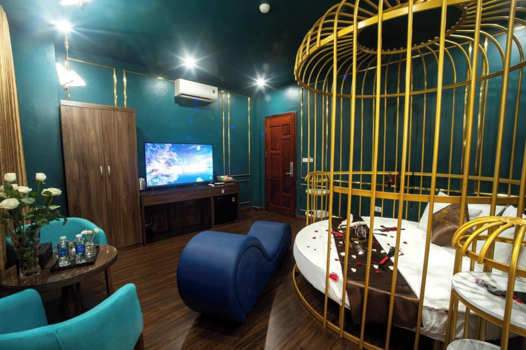 A room with a TV, wooden furniture, a blue sofa, a bed with a golden cage, and a small table with a vase and bottles.