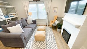 Una sala de estar con un sofá gris, otomanas, un tapete estampado, una chimenea y un televisor montado en la pared.