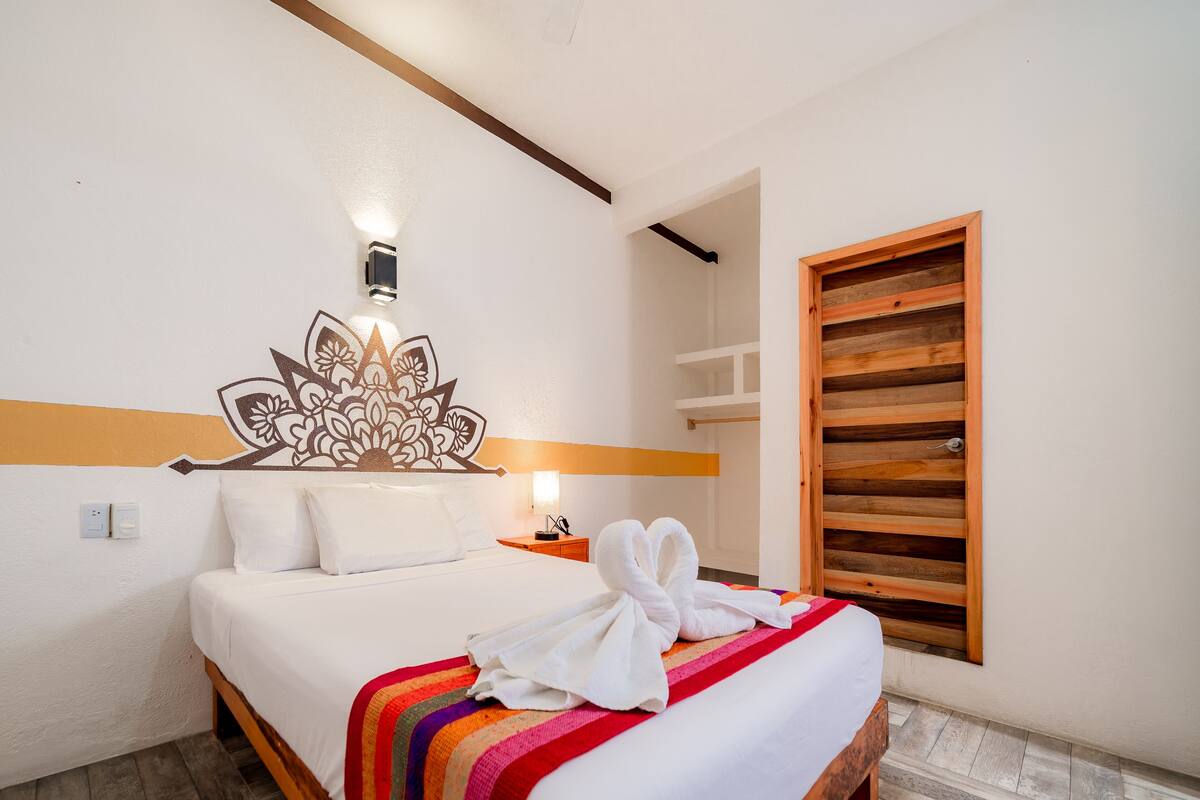 Une chambre d’hôtel avec un lit, une lampe fixée au mur, une tête de lit en bois et une fenêtre avec des stores.