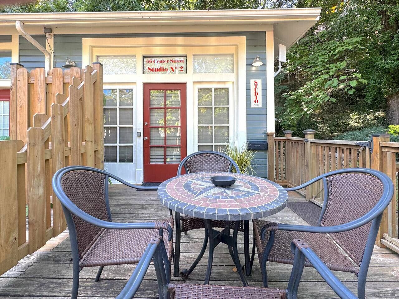 A patio with a round table and four wicker chairs, a wooden fence, and a building with a red door and a sign reading "66 Center Street Studio No. 2".