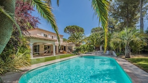 Ein Schwimmbecken mit klarem, blauem Wasser, umgeben von üppiger Vegetation und einer Villa mit einem Ziegeldach.