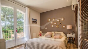Ein Schlafzimmer mit einem großen Bett, einem Nachttisch, einer Lampe und einer Wanddekoration. Ein Fenster mit Vorhängen bietet Blick auf Grünflächen.