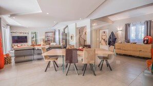 Ein geräumiger Essbereich mit großem Holztisch und modernen Stühlen, eine Küche mit Einbauofen sowie ein Wohnbereich mit Sofa und Fernseher.