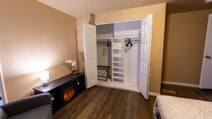 Quarto de hotel com cama, área de estar com luminária, pequena mesa com planta e armário com portas abertas.
