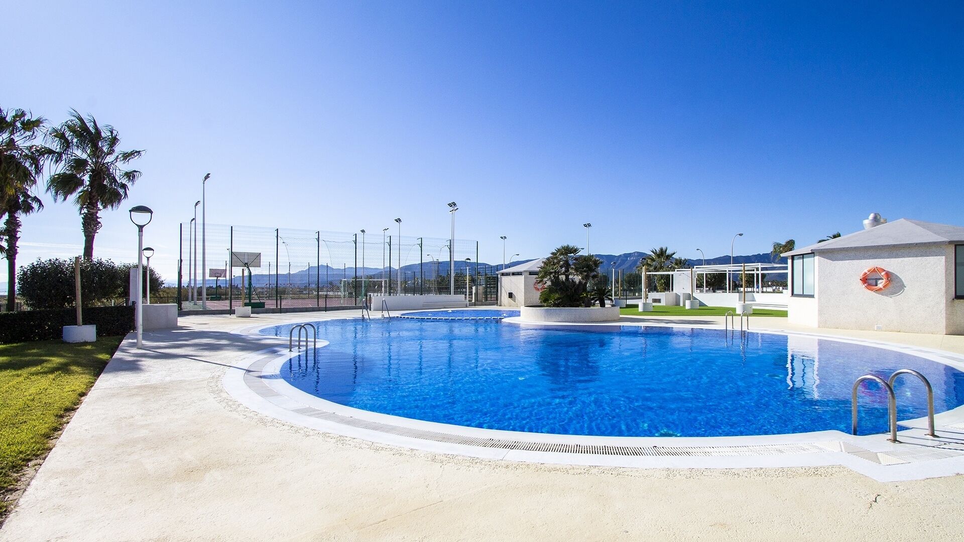 Una grande piscina trasparente con una passeggiata circostante, un campo da basket e un salvagente appeso a una parete dell'edificio.