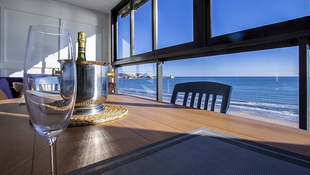 Ett bord med ett glas vatten, en flaska champagne och en underlägg. Bordet står nära ett fönster med utsikt över havet, och det finns en stol i närheten.