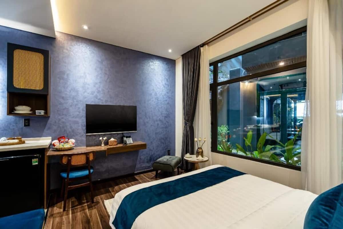 A hotel room with a bed, a desk with a TV, a small table, and a window with a view of greenery.