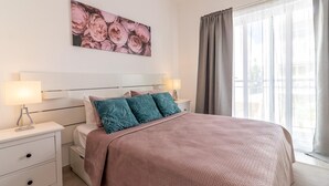 Een slaapkamer met een groot bed, twee nachtkastjes met lampen, een bloemstillevoorstelling aan de muur, en uitzicht door een raam met gordijnen.
