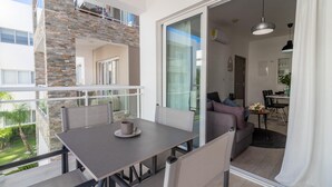 Een balkon met een tafel en stoelen, een zithoek met een bank en uitzicht op een tuin en gebouw.