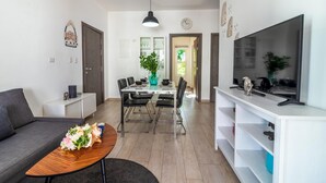 Een moderne woonkamer met een bank, een salontafel met bloemen, een eethoek met een tafel en stoelen, een tv op een dressoir en een gang die leidt naar een deur.