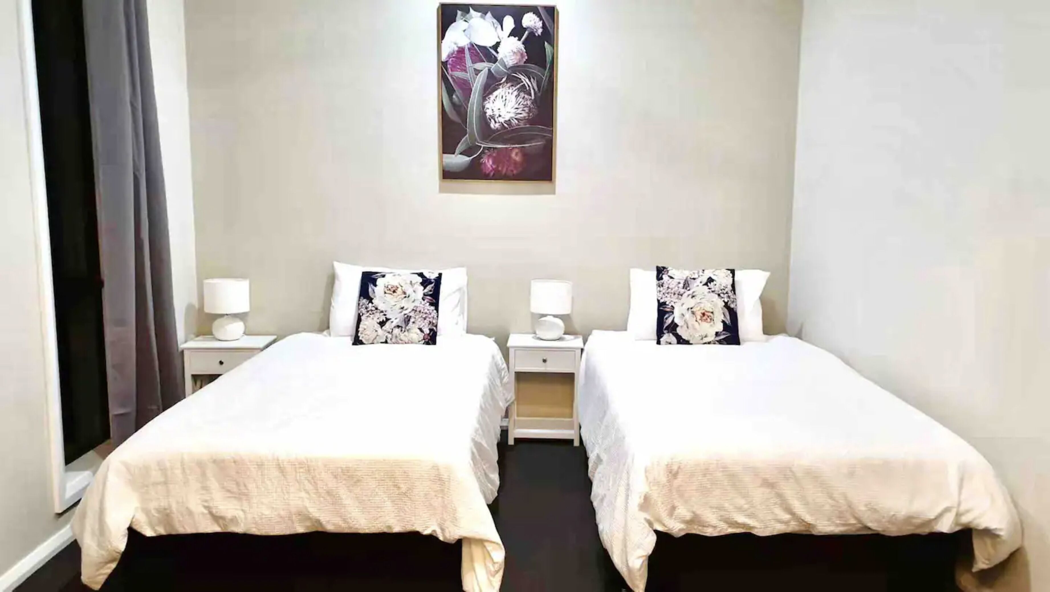 Ett hotellrum med två sängar, var och en med ett vitt överkast och en prydnadskudde med blommotiv. Vid sängarna står nattduksbord med lampor, och på väggen hänger en tavla med blommor.