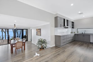 A modern kitchen with grey cabinets, a white countertop, and a built-in oven. There is a dining area with a wooden table and chairs, and a potted plant.