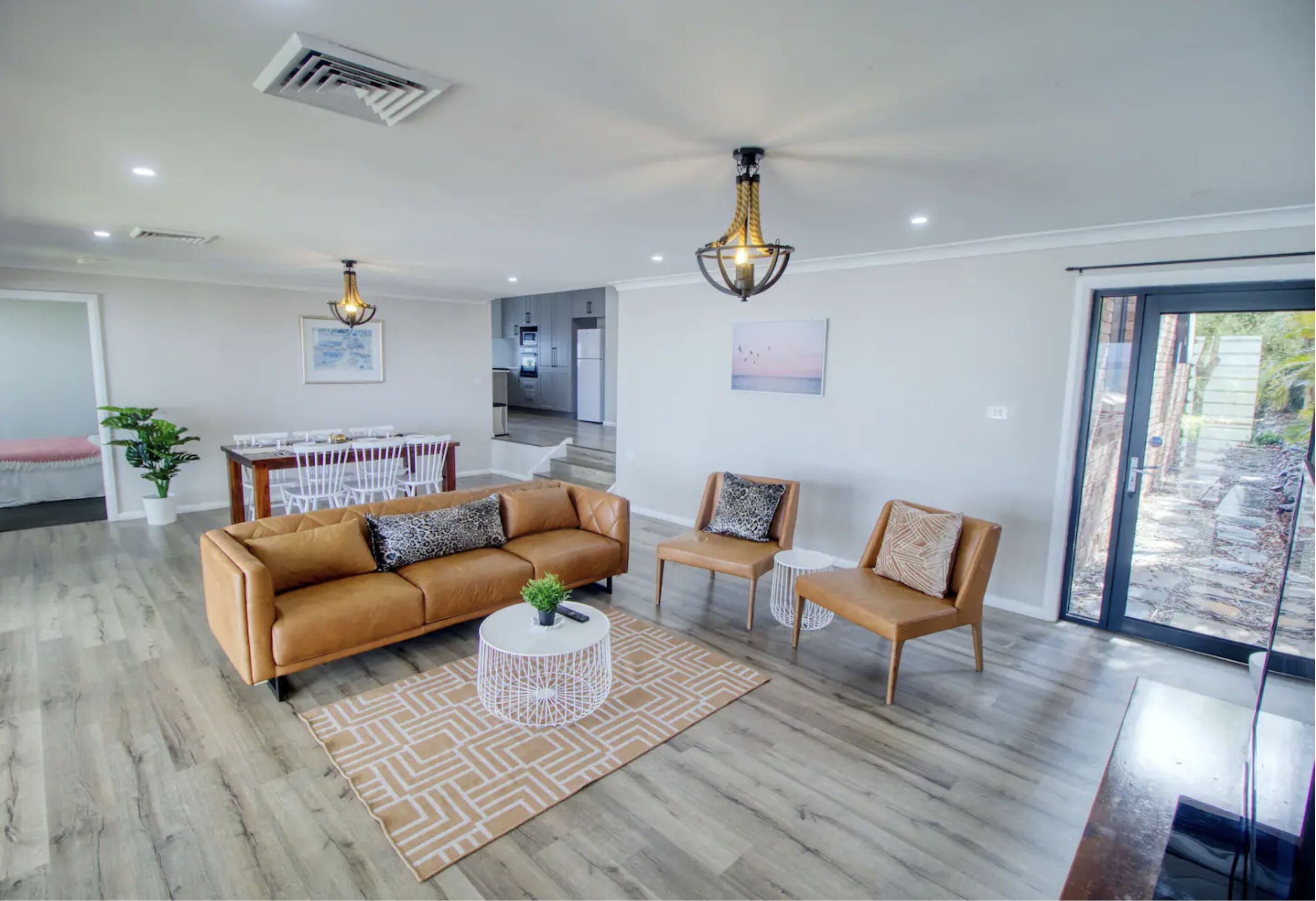 A modern living room with a sofa, chairs, and a coffee table. There is a dining area with a table and chairs. The room has a large window and a view of the outside.