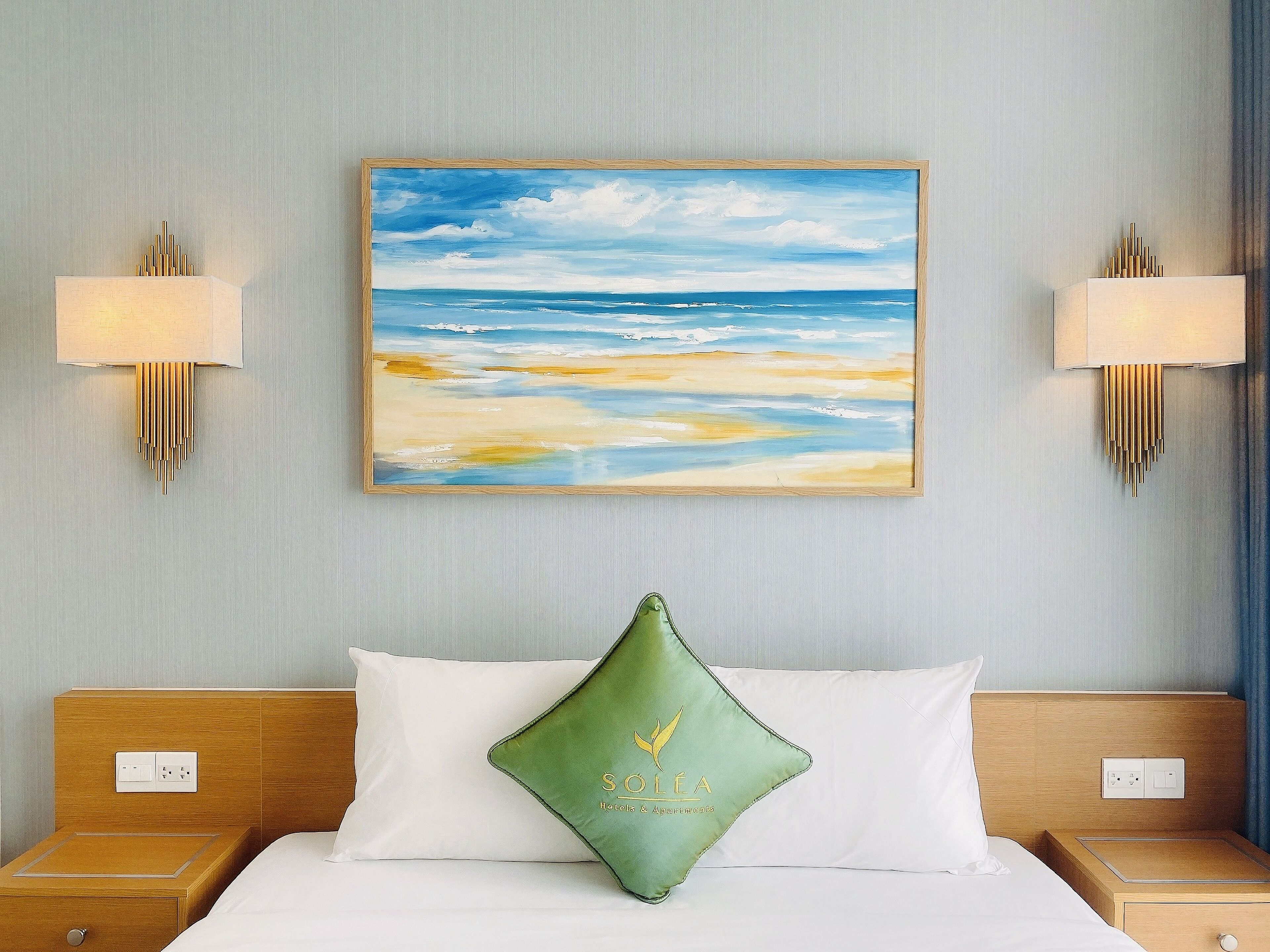 Una habitació d'hotel amb un llit, un coixí verd, un quadre de platja i làmpades penjades a la paret.