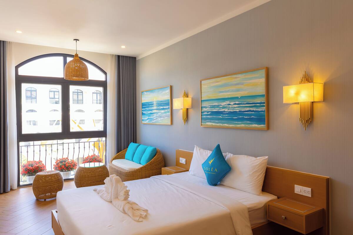 A hotel room with a large bed, a wicker chair, a nightstand, and a view of the city through a large window.