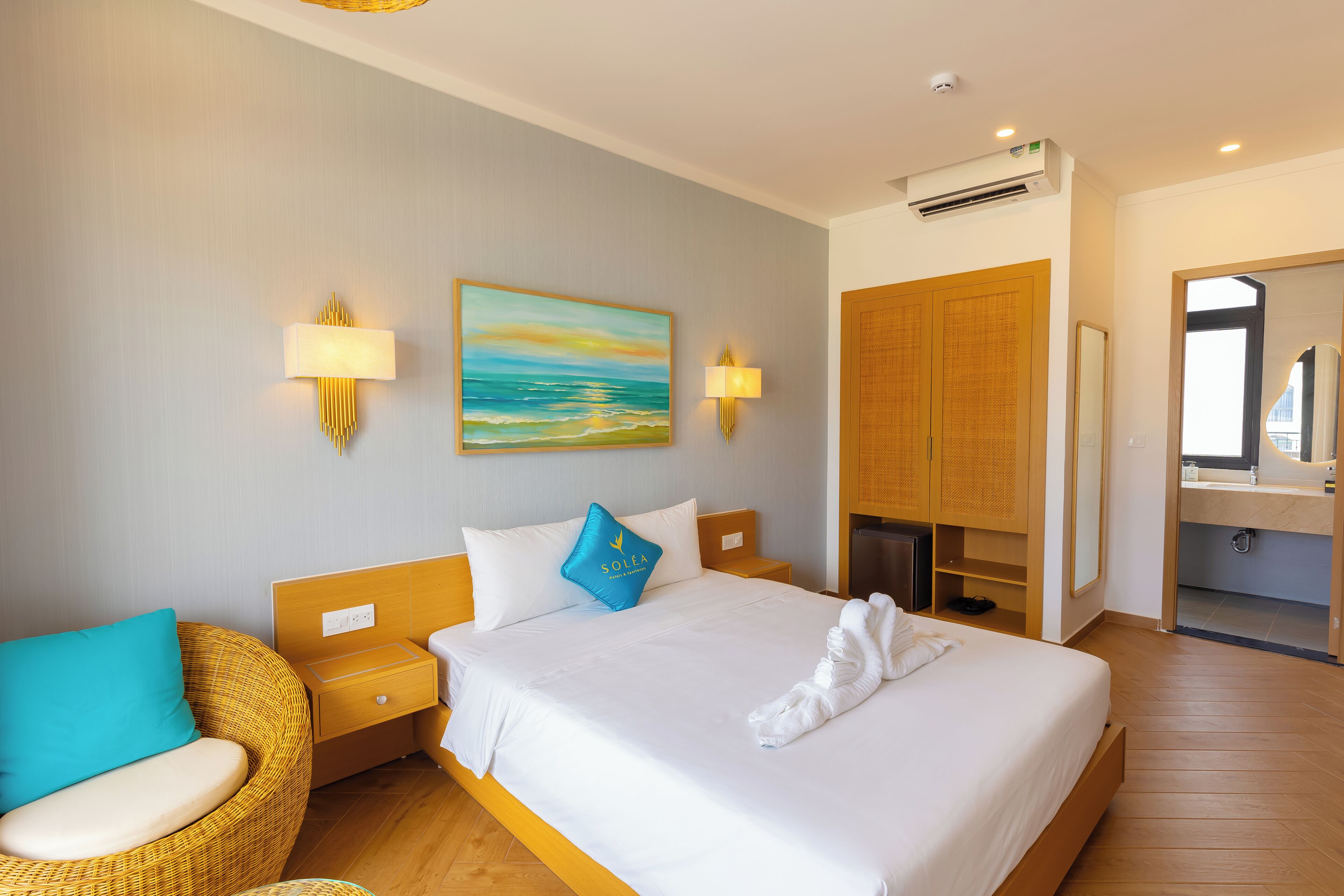 Habitación de hotel con una cama, una mesita de noche, un sillón de mimbre, un cuadro, un armario y un baño visible a través de una puerta abierta.