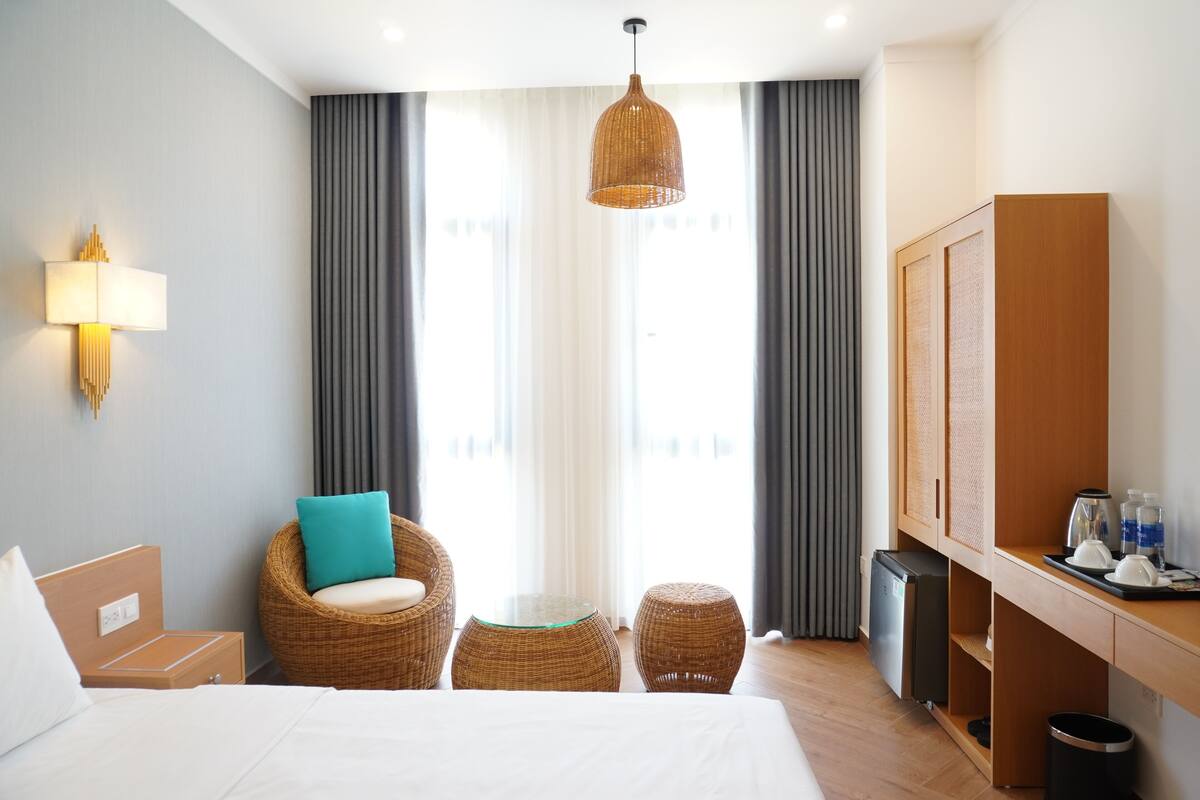 A hotel room with a bed, a wicker chair with a teal cushion, a small table, a television, and a minibar.