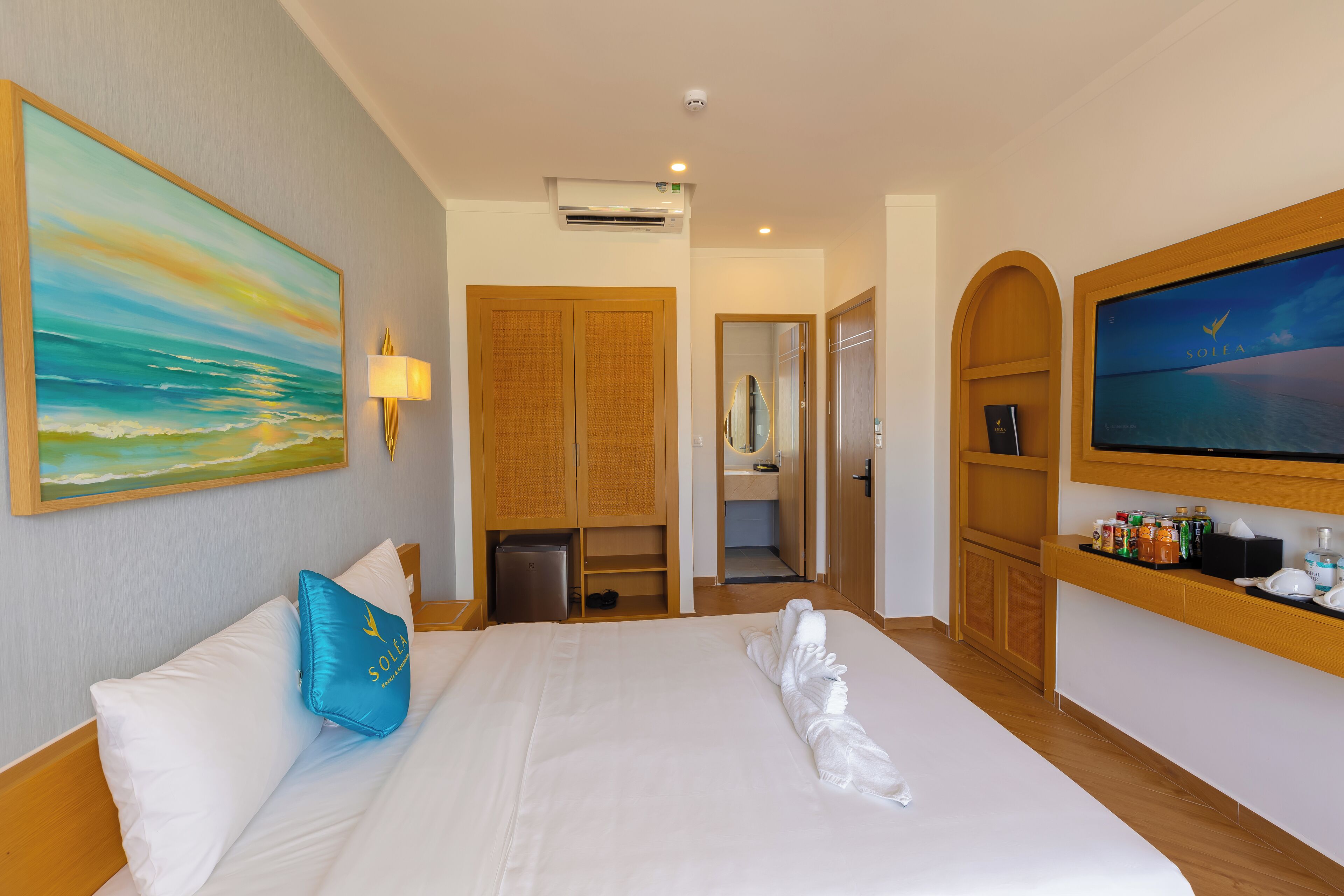 A hotel room with a bed, a painting, a TV, and a minibar.