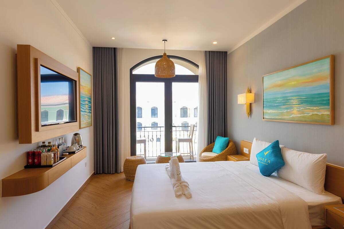 A hotel room with a large bed, a television, a balcony with a view of buildings, and a dining area with wicker furniture.