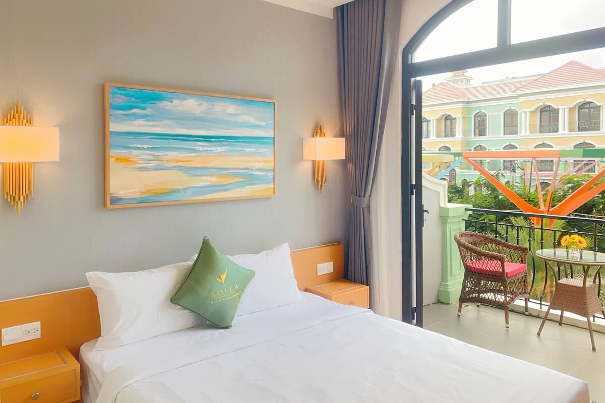 A hotel room with a bed, a painting of a beach, a balcony with wicker furniture, and a view of buildings.