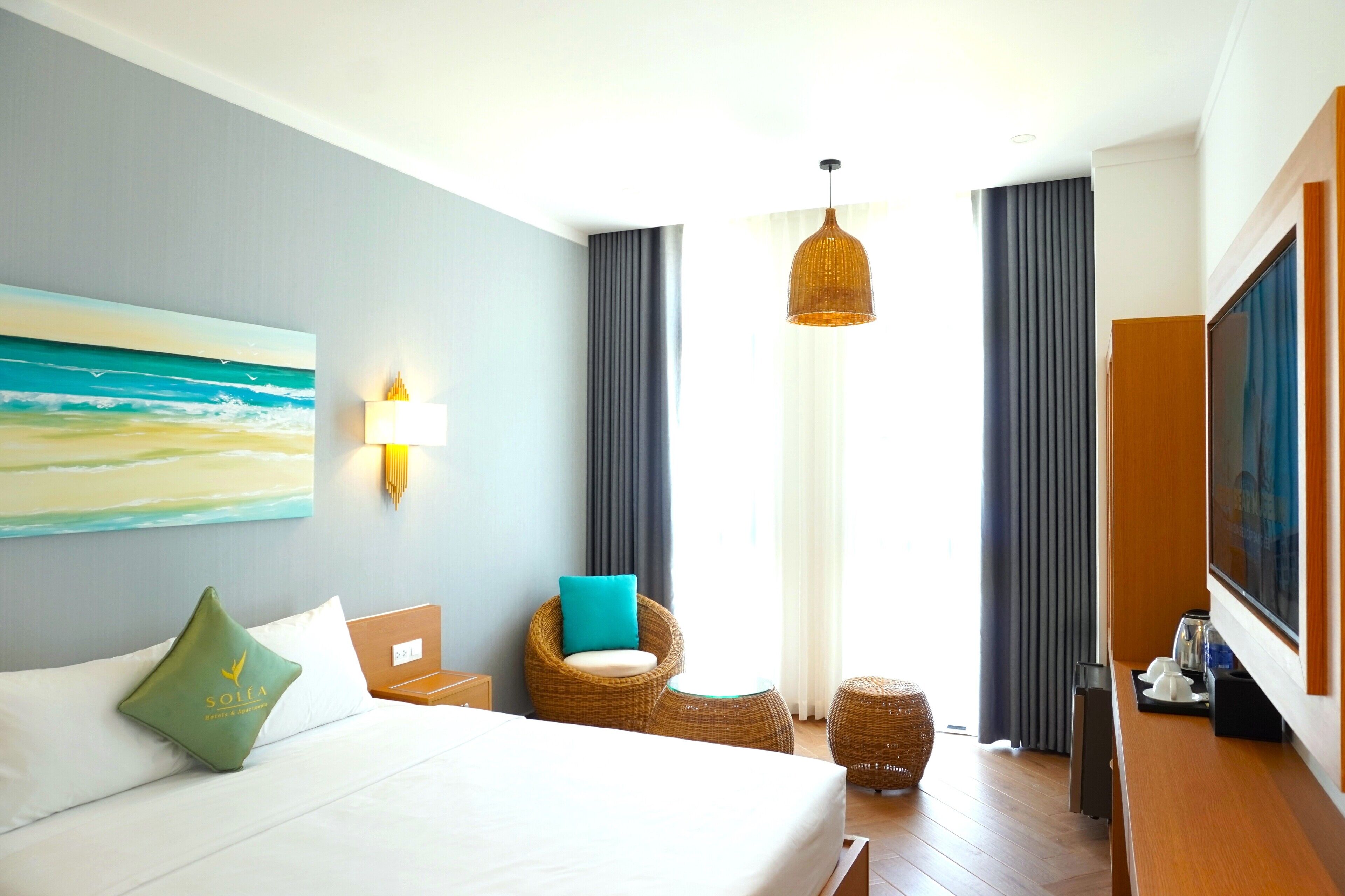Une chambre d’hôtel avec un lit, une chaise en osier, une petite table ronde, un téléviseur et un tableau au mur.
