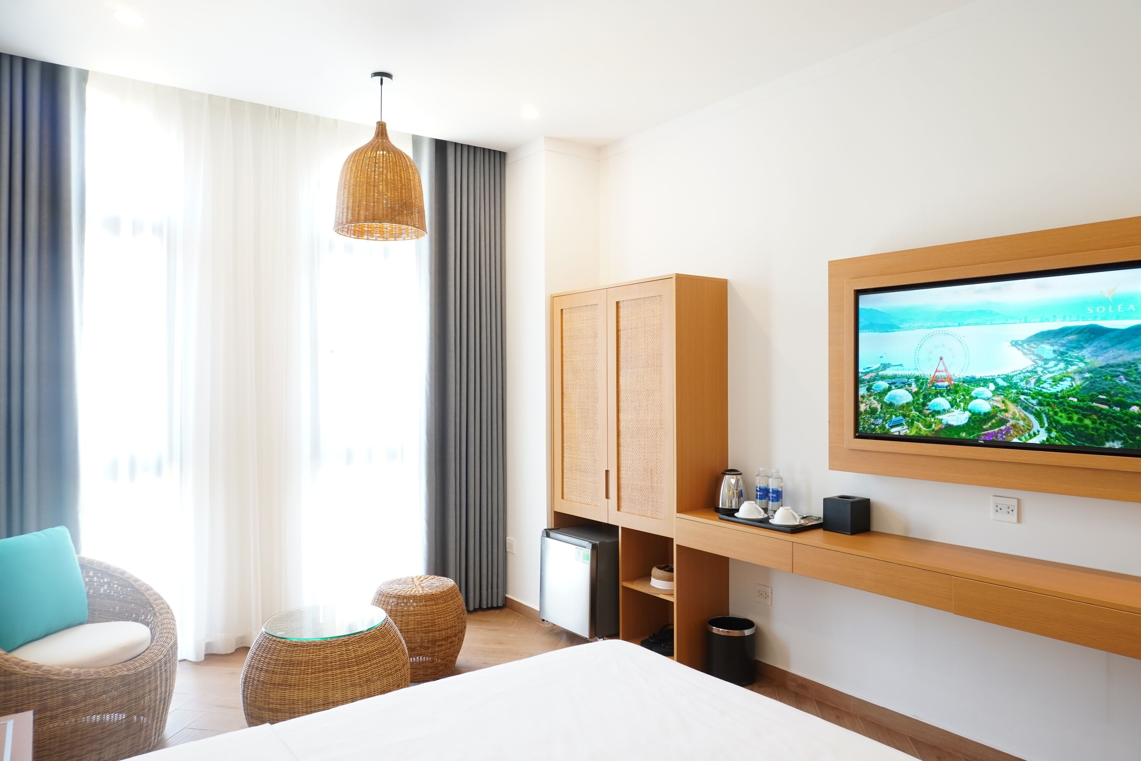 Een hotelkamer met een bed, een aan de muur bevestigde televisie, een kleine koelkast, een rieten stoel en een raam met gordijnen.