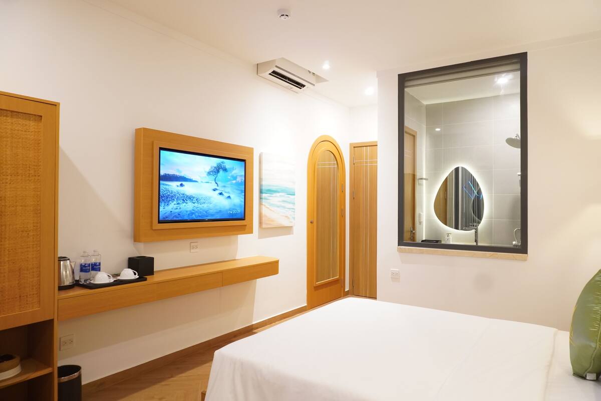 A hotel room with a bed, a mounted TV, a wooden shelf with toiletries, a mirror, and a door.