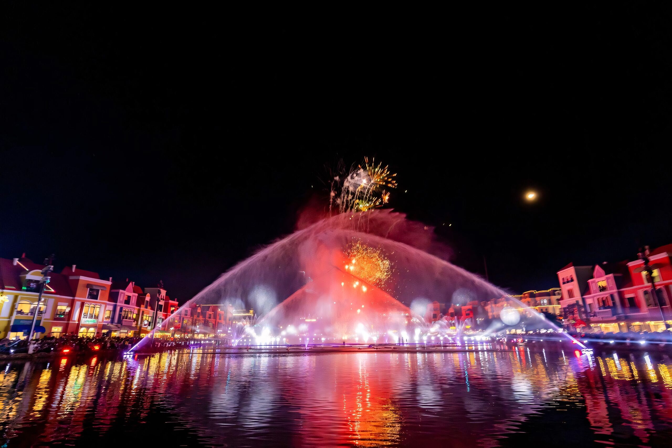 Vista nocturna de una fuente de agua con luces de colores y fuegos artificiales, reflejados en el agua, con edificios al fondo.