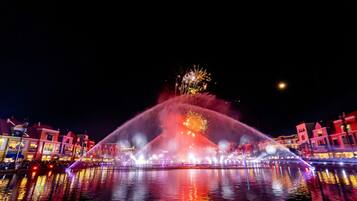 Vista nocturna de una fuente de agua con luces de colores y fuegos artificiales, reflejados en el agua, con edificios al fondo.